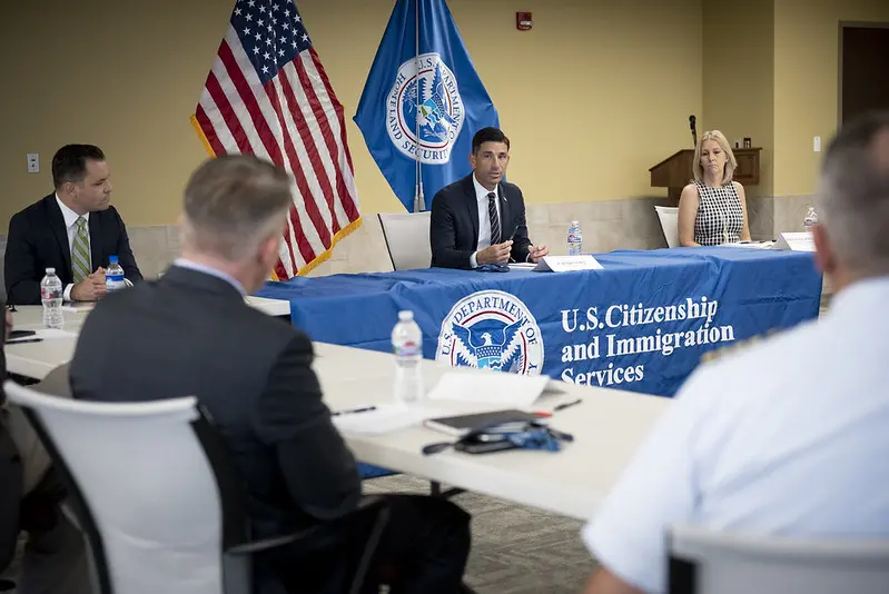 A group of people sit around a conference table at a U.S. Citizenship and Immigration Services meeting, with USCIS banners and American flags in the background. One man speaks while others listen attentively.