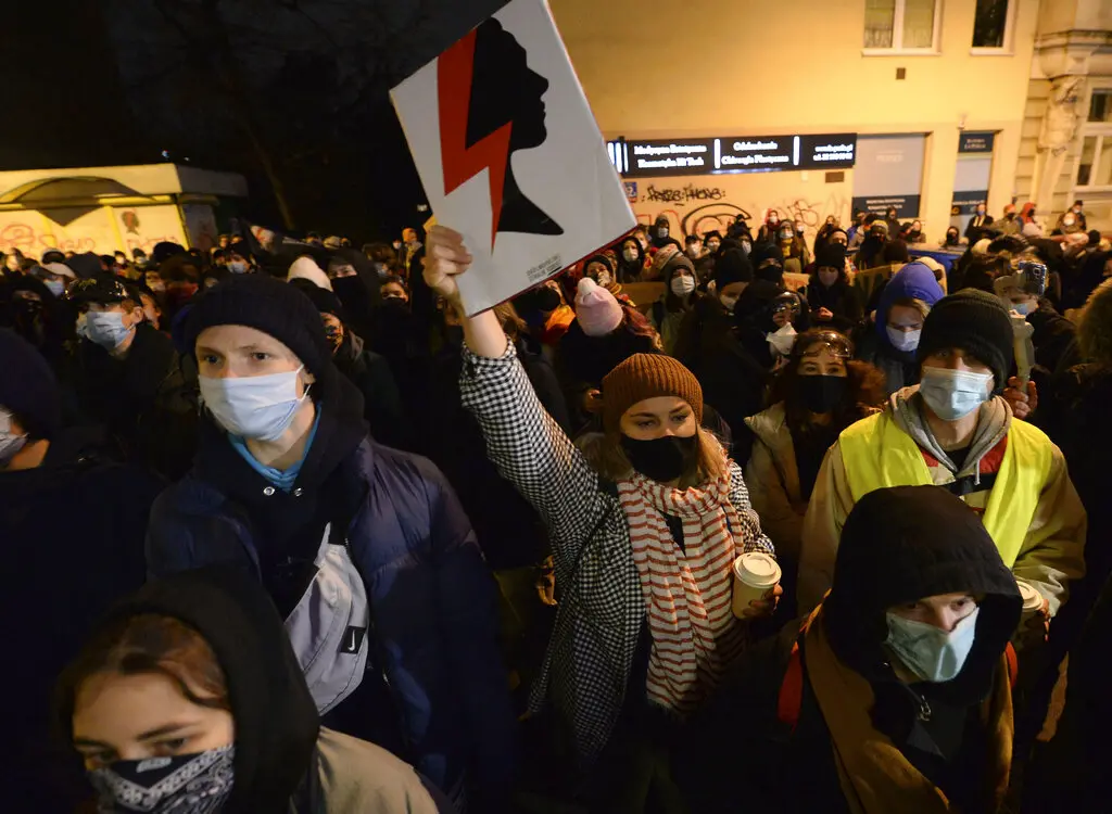 A large crowd of people, many wearing masks, gathers outdoors. A person in the center holds up a sign with a silhouette and a red lightning bolt. It appears to be a protest or demonstration at night.