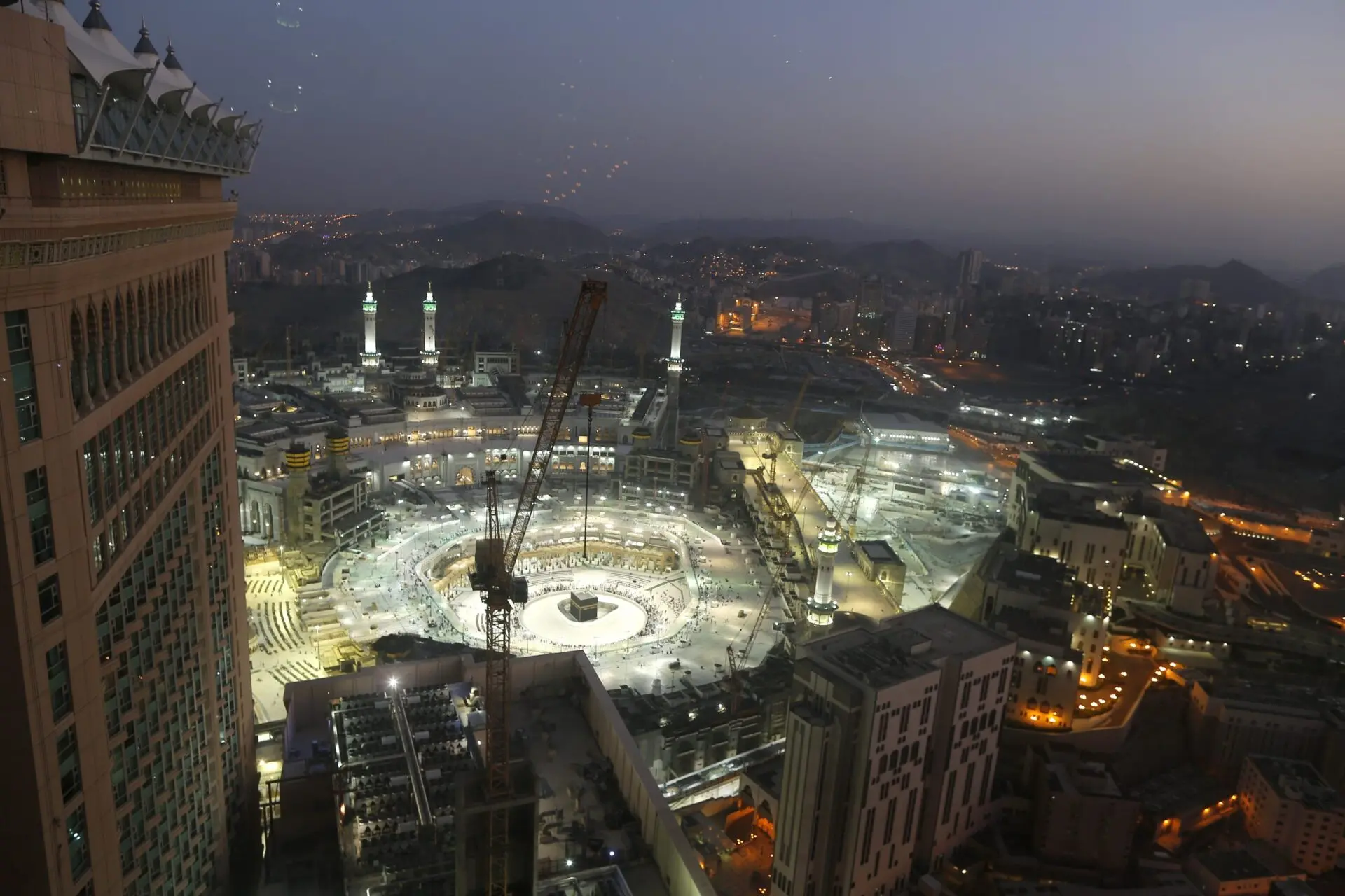 Aerial view of the Grand Mosque in Mecca at dusk, brightly illuminated and surrounded by city buildings, with construction cranes visible and distant hills in the background.