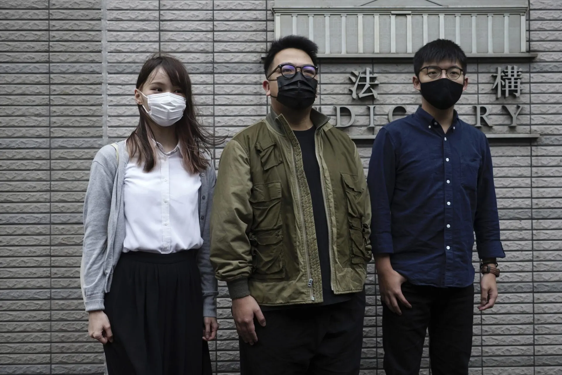 Three people wearing face masks stand side by side in front of a gray brick wall with a sign in the background. They appear to be posing for the photo, dressed in casual and semi-formal clothing.