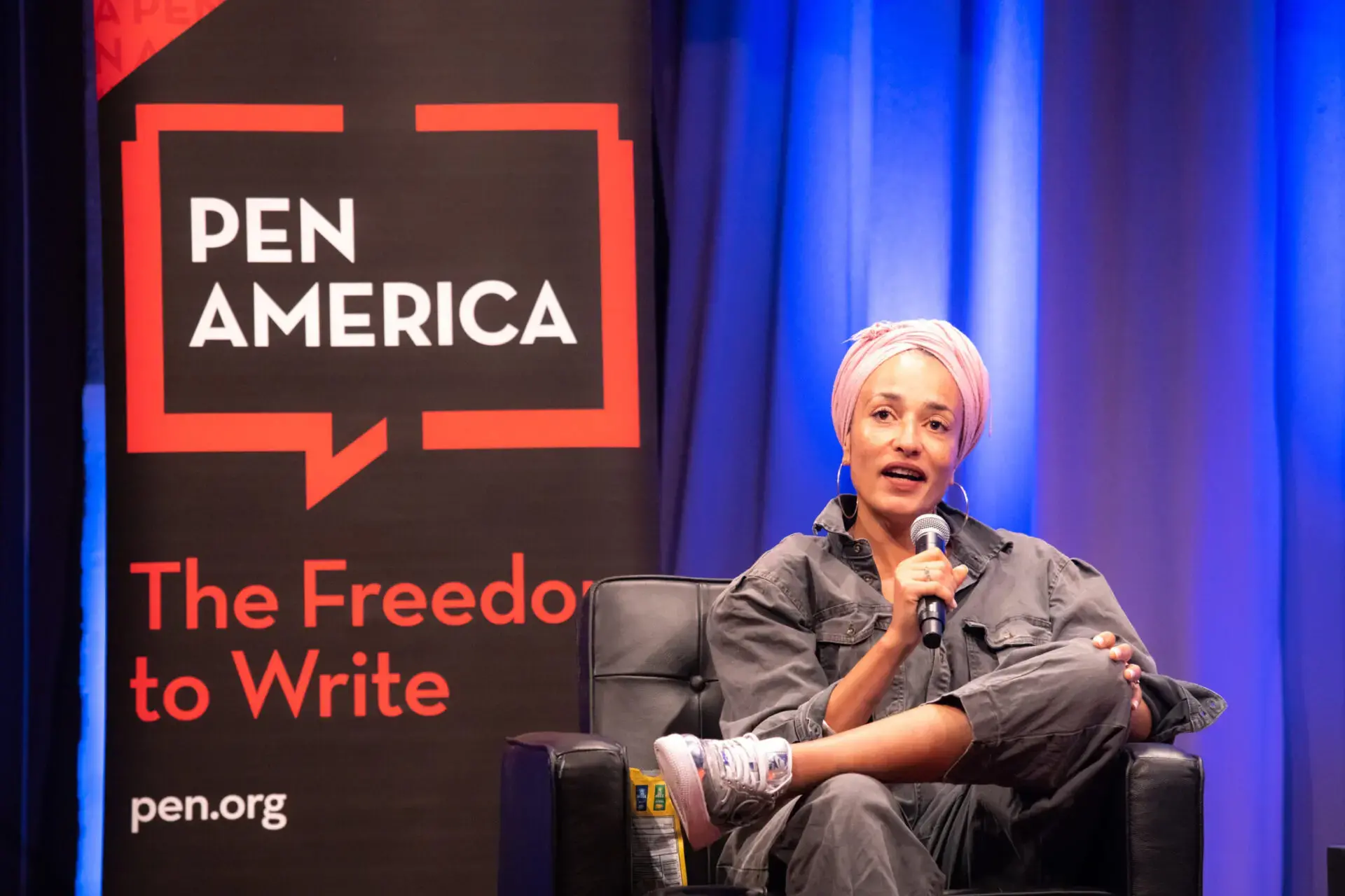 A person in a gray outfit and pink headwrap speaks into a microphone while sitting on stage beside a black PEN America banner that reads, “The Freedom to Write,” during a PEN America news event.