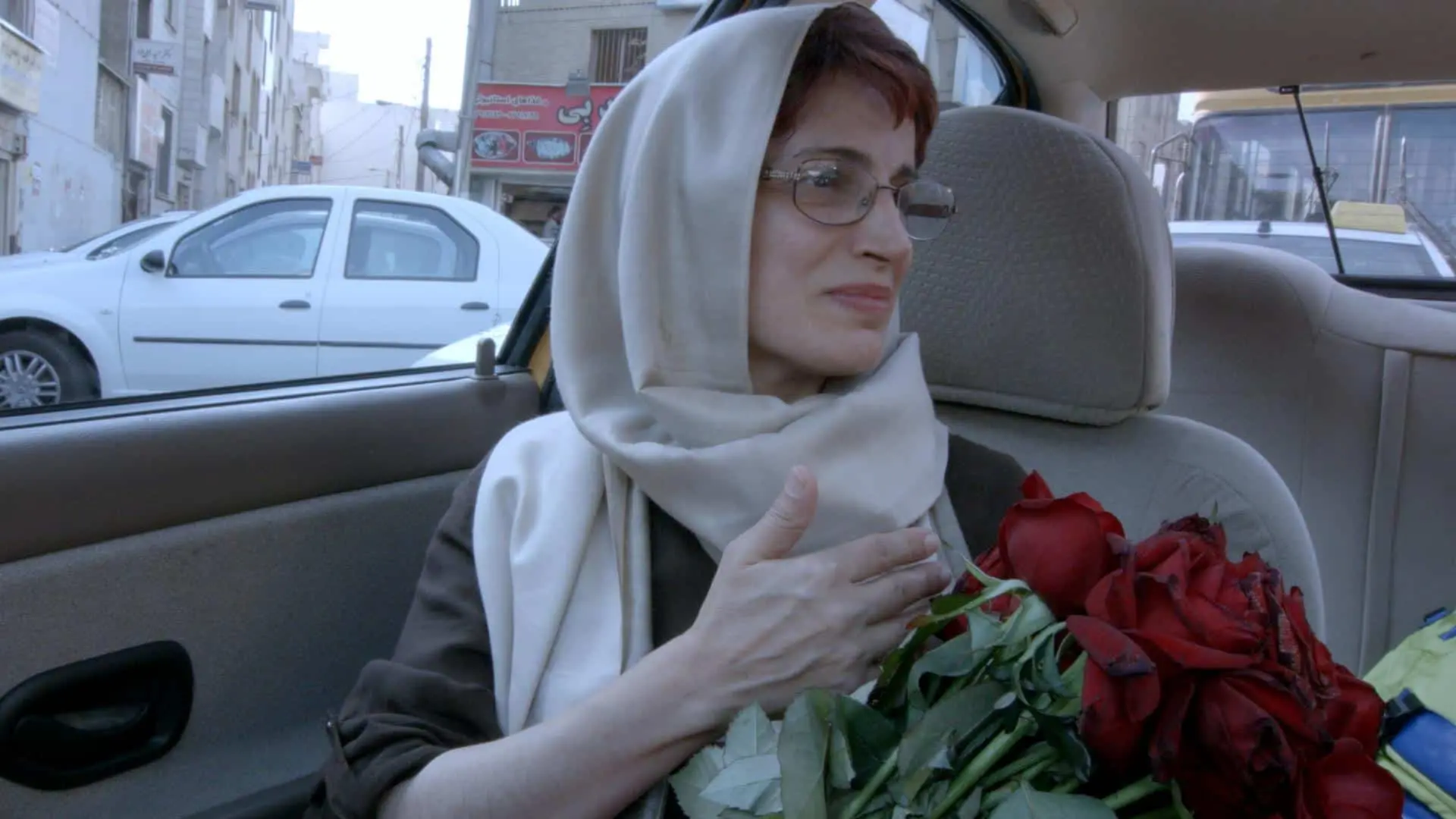 A woman wearing glasses and a light-colored headscarf sits in a car, holding a bouquet of red roses and looking out the window with a gentle expression. Urban buildings and a white car are visible outside.