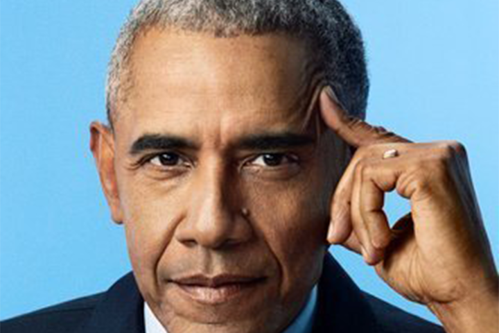 A man with short gray hair wearing a dark suit poses against a blue background, looking directly at the camera with a thoughtful expression and his index finger resting on his temple at the PEN America Literary Gala.
