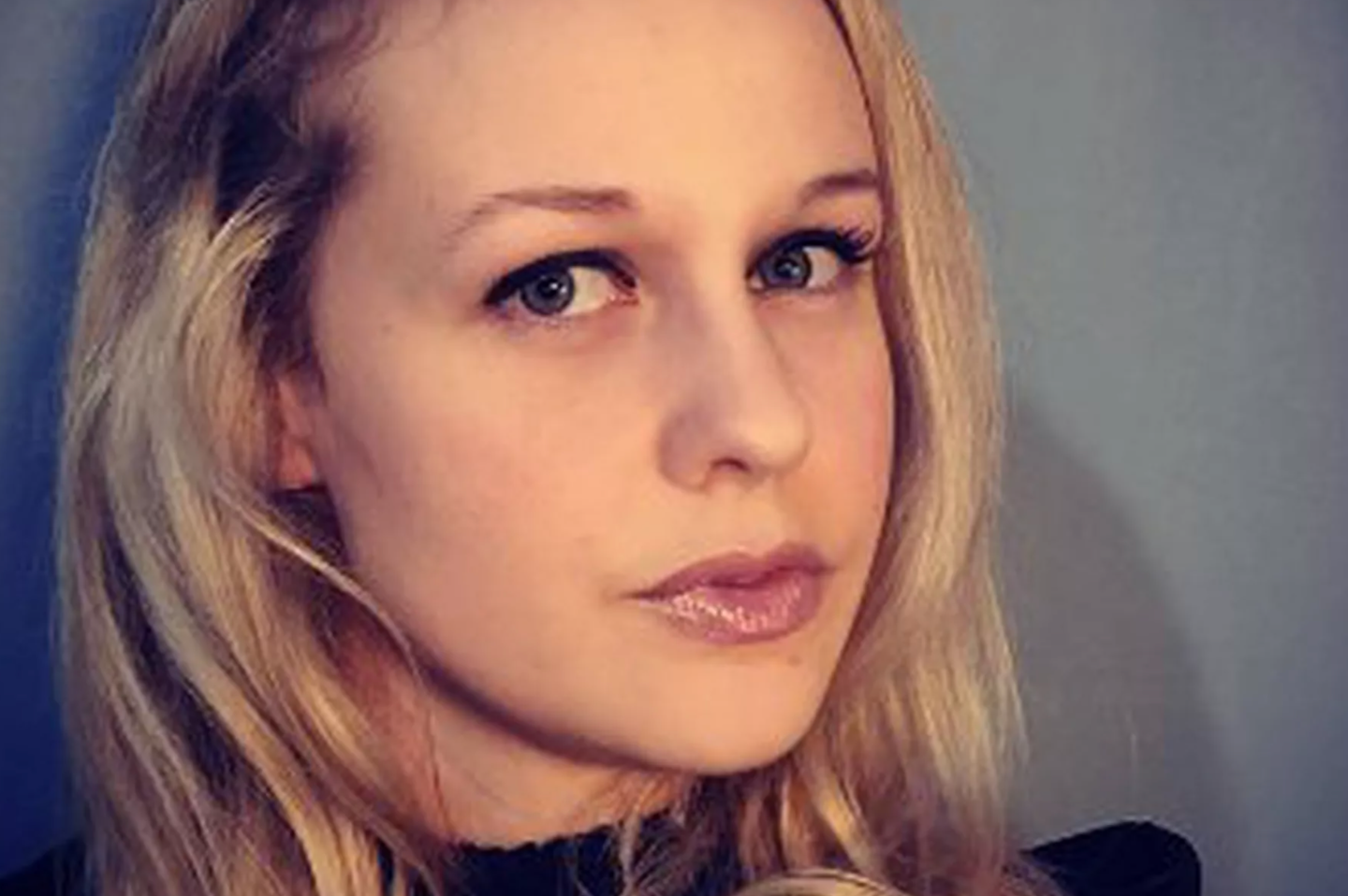 A woman with long blonde hair and light eyes looks into the camera with a neutral expression. She is wearing light makeup and a dark top, with a simple background behind her.