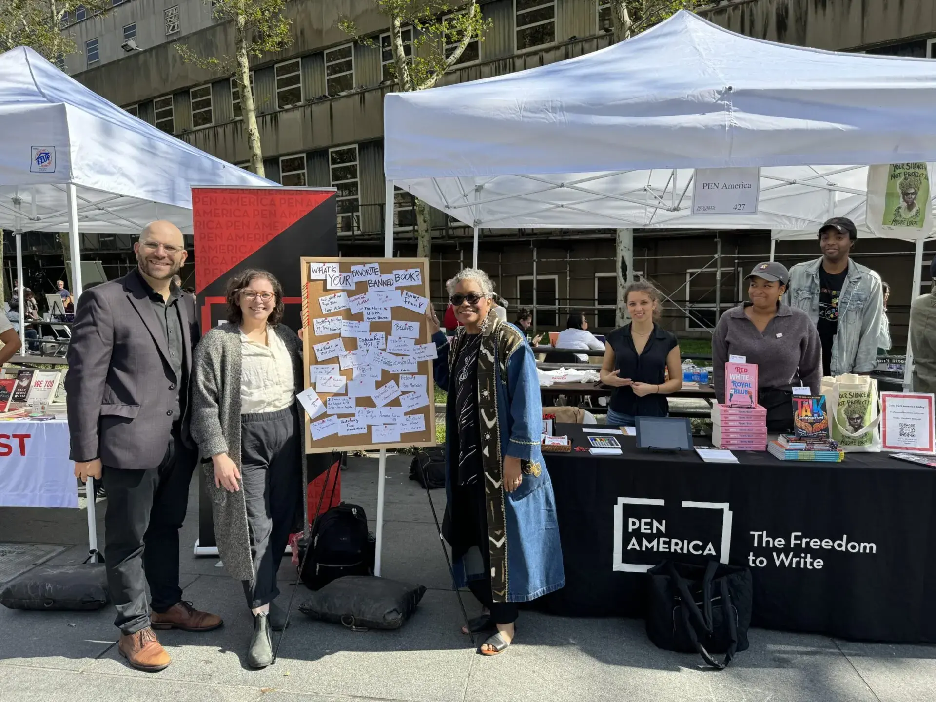 A group of people stand and sit at an outdoor booth for PEN America, featuring books, flyers, a board covered in handwritten notes, and updates on PEN America news, with tents and buildings in the background.
