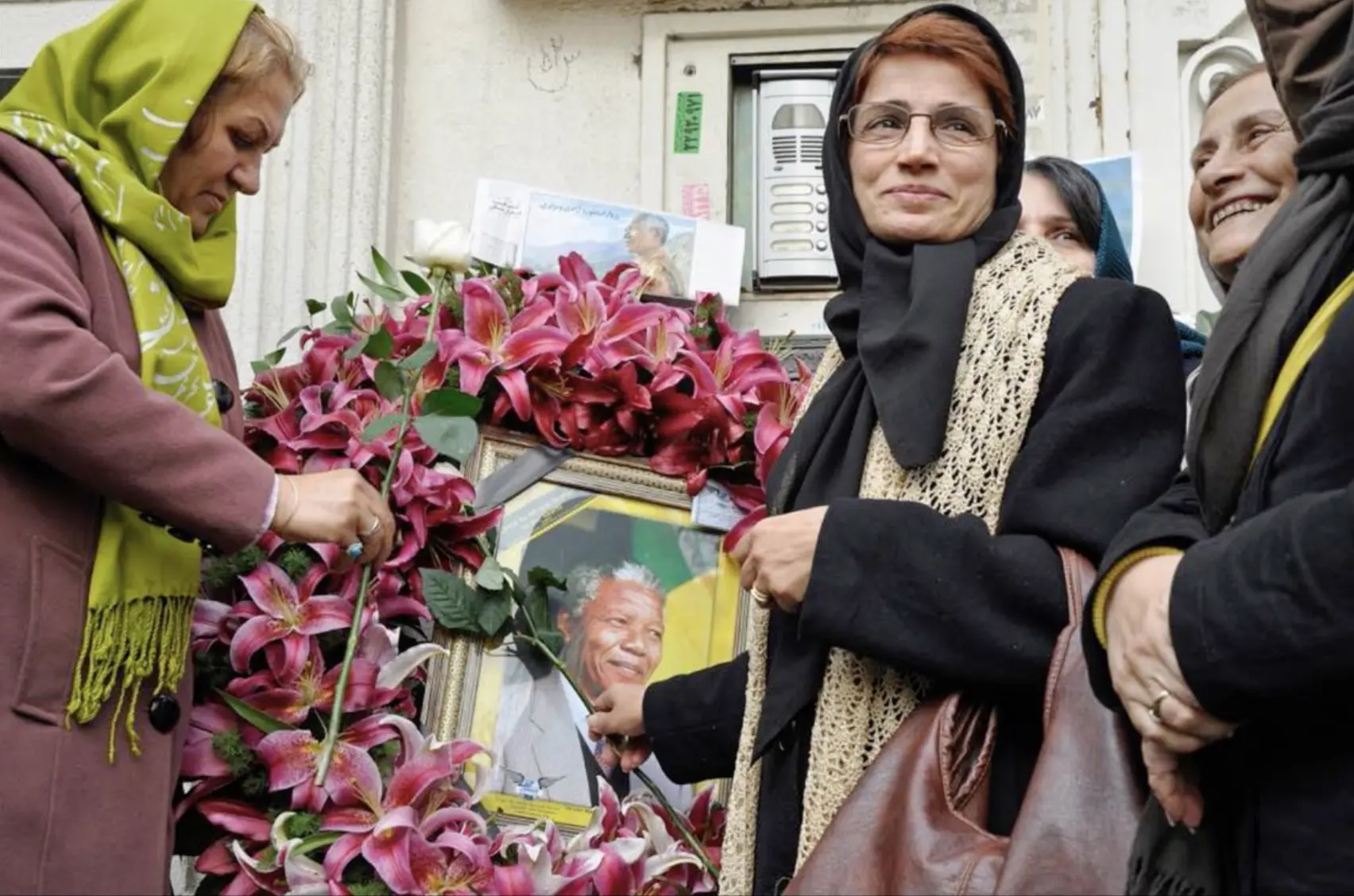Three women in headscarves stand by a large wreath of pink lilies surrounding a framed photo of Nelson Mandela, appearing to honor or pay tribute to him. One woman smiles while holding the frame.