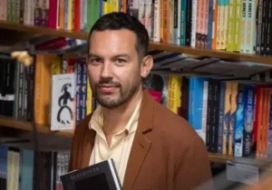 A person with short dark hair and a beard, wearing a brown blazer and holding a book, stands in front of shelves filled with colorful books, possibly browsing the latest PEN America news in a bookstore or library.