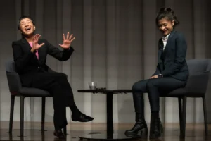 Two women in suits sit on stage in conversation at a PEN America news event, one laughing with hands raised, the other smiling with hands in her lap. A small table with two glasses of water sits between them.