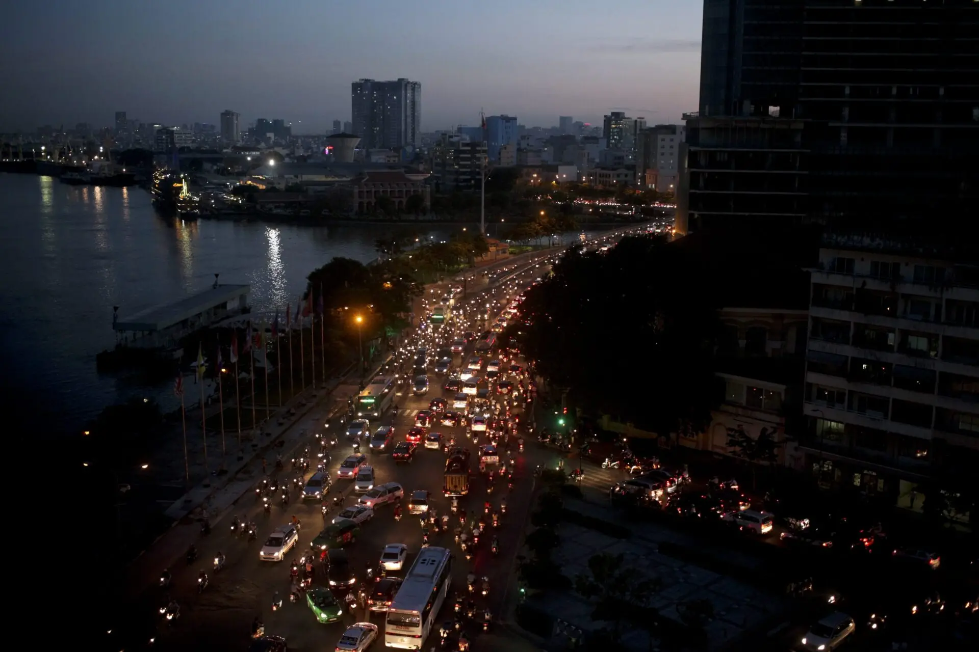A busy city street at dusk, filled with heavy traffic of cars and motorcycles. Lights from vehicles and buildings illuminate the scene, with a river and high-rise buildings visible in the background.