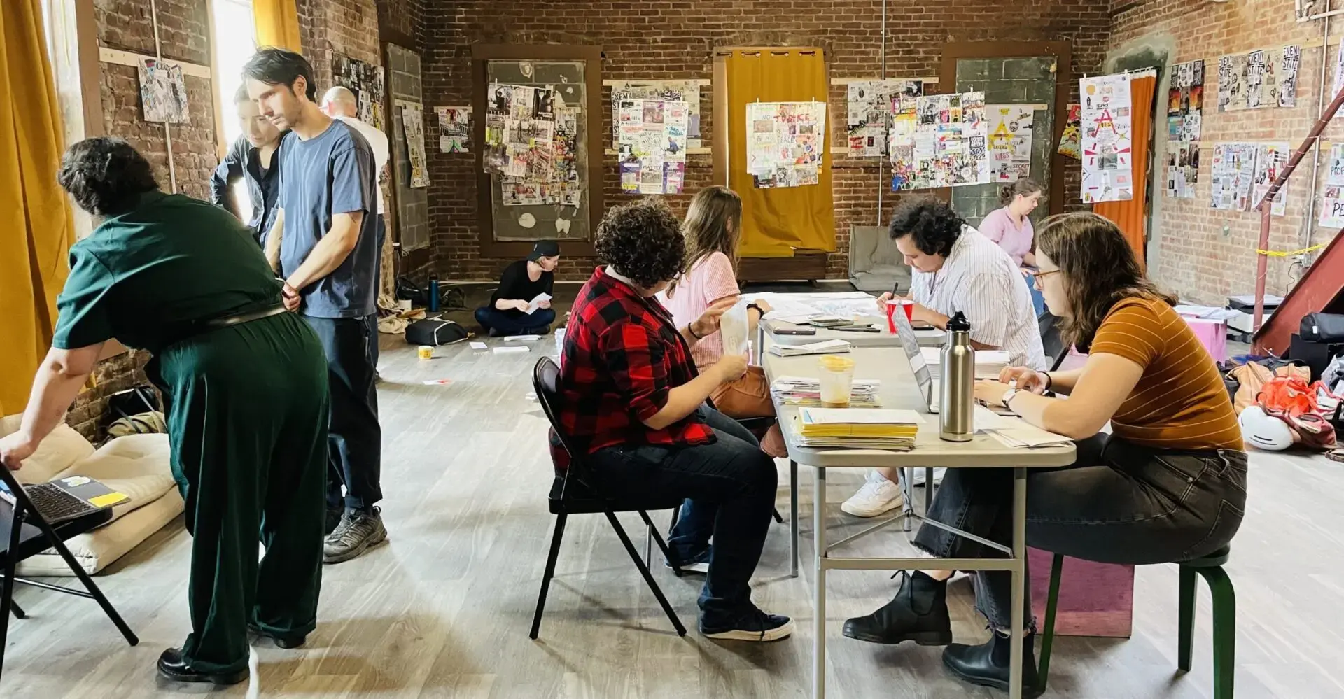 A group of people work and interact in a casual, creative studio with exposed brick walls, posters, and papers about Prison Library Support Network reference letters on the walls. Some sit at a table with laptops and drinks; others converse on the floor or standing.