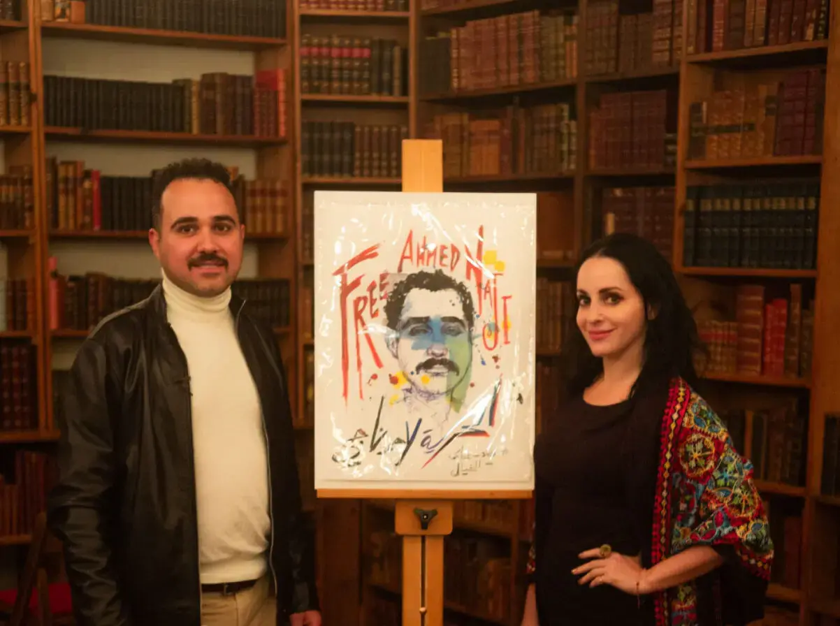 Two people stand on either side of an easel holding a colorful portrait with the text “Free Ahmed.” They are in a library full of books, highlighting PEN America news and advocacy for writers’ rights.