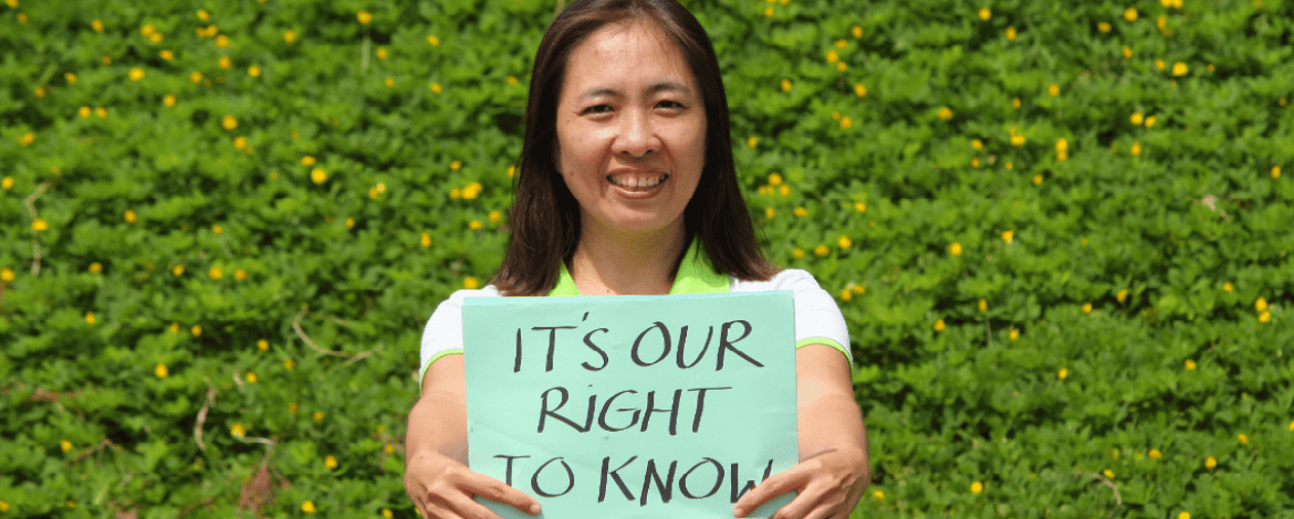 A woman smiling outdoors holds a sign that reads, ITS OUR RIGHT TO KNOW, with green foliage and yellow flowers in the background.