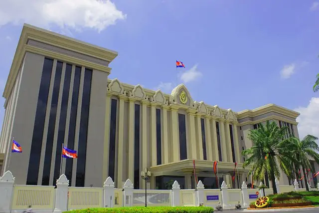 A large government building with columns, a triangular pediment, and a clock above the entrance, with Cambodian flags on the roof and grounds, surrounded by a white fence and palm trees under a blue sky.