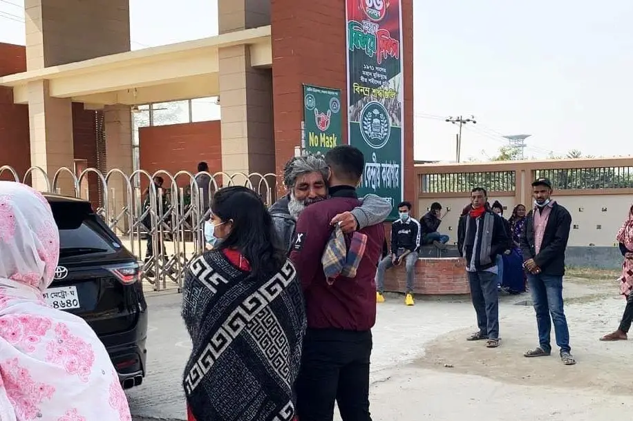 A man and a young person hug outside a building with a sign that reads No Mask No Entry. Several people stand or walk nearby, some wearing masks, and cars are parked along the side.