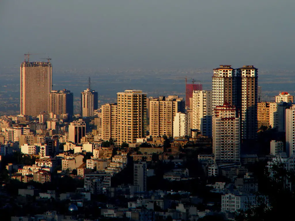 A cityscape at sunset showing numerous tall buildings and skyscrapers, some under construction, with shadows and sunlight highlighting the structures against a hazy, distant horizon.