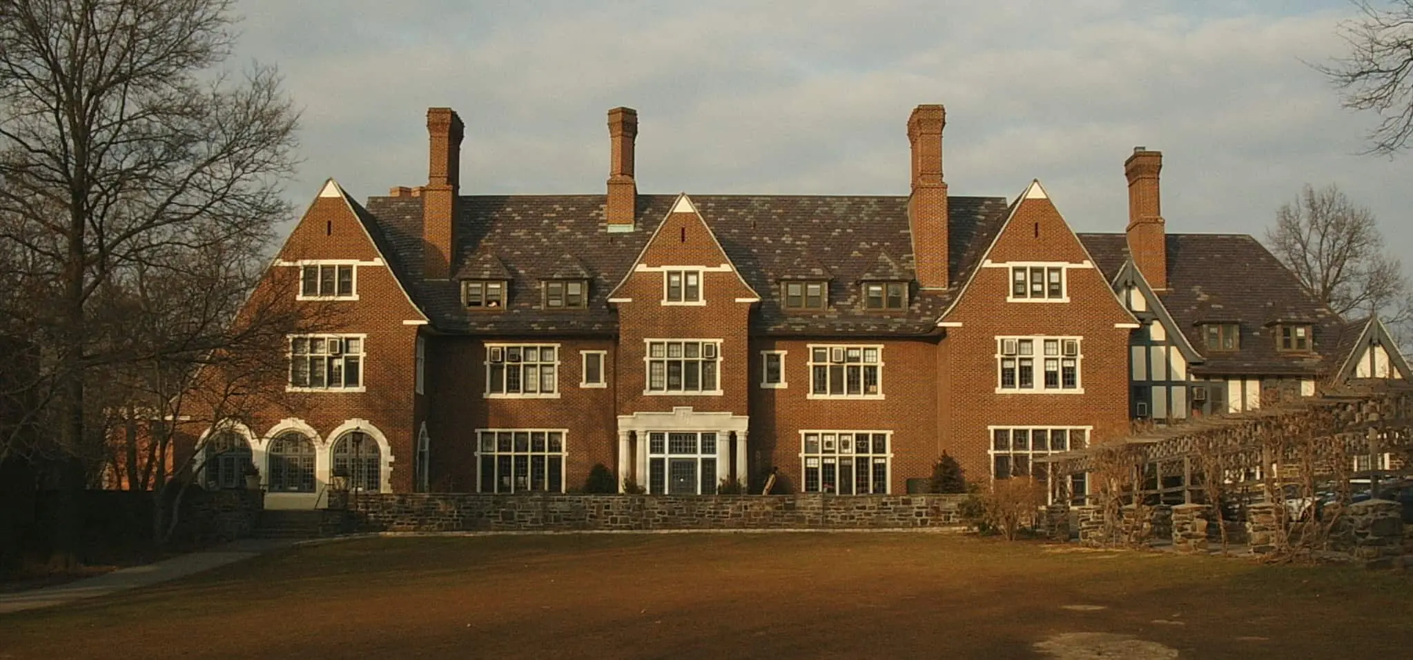 A large, historic brick mansion with multiple chimneys, tall windows, and a stone foundation sits in front of a lawn under a partly cloudy sky. Leafless trees frame the scene.