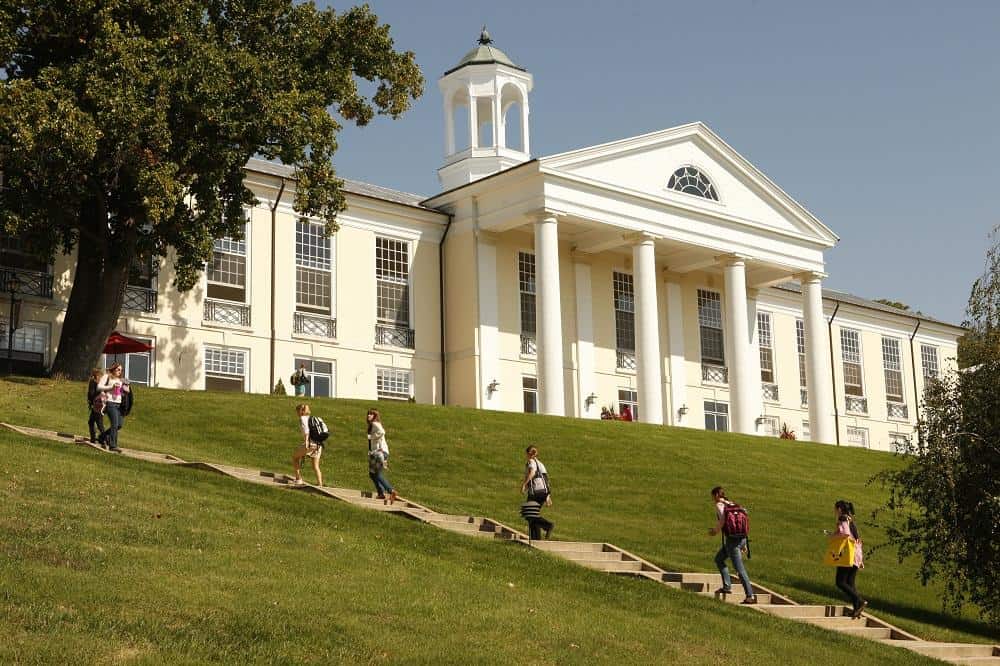 Students walk up and down a sloped pathway in front of a large, white, neoclassical building with columns and a cupola, surrounded by green grass and trees on a sunny day.