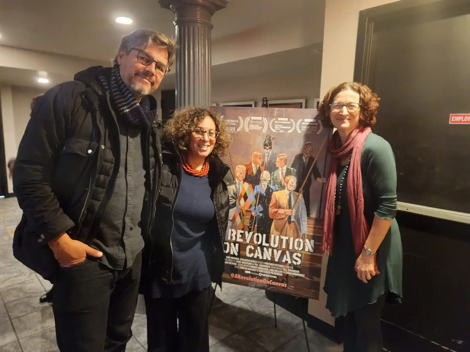 Three people stand smiling beside a colorful “Revolution on Canvas” movie poster in an indoor setting with dim lighting and tiled floors, as featured in recent PEN America news. The poster displays illustrated figures and awards laurels at the top.