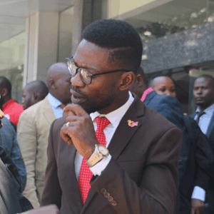 A man in a brown suit, red tie, and glasses stands outdoors among a group of people, thoughtfully touching his chin.