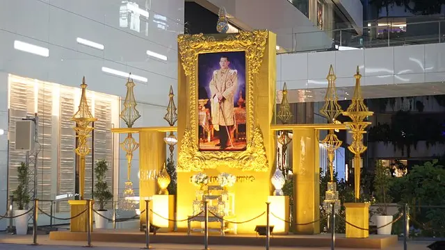 A large, ornate golden frame displays a royal portrait of a person in formal attire, set within an illuminated, gold-themed shrine in a modern indoor space, surrounded by decorative pillars and roped barriers.