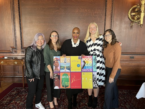 Five women, including Ayanna Pressley, stand together smiling in a wood-paneled room. The woman in the center holds a collage of colorful banned book covers, like Gender Queer, Lawn Boy, and This Book is Gay.