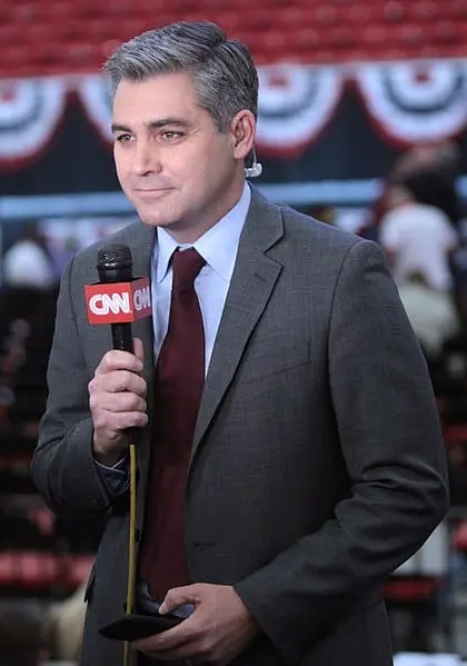 A man in a gray suit and maroon tie holds a CNN microphone while reporting indoors, with festive red, white, and blue bunting in the blurred background.