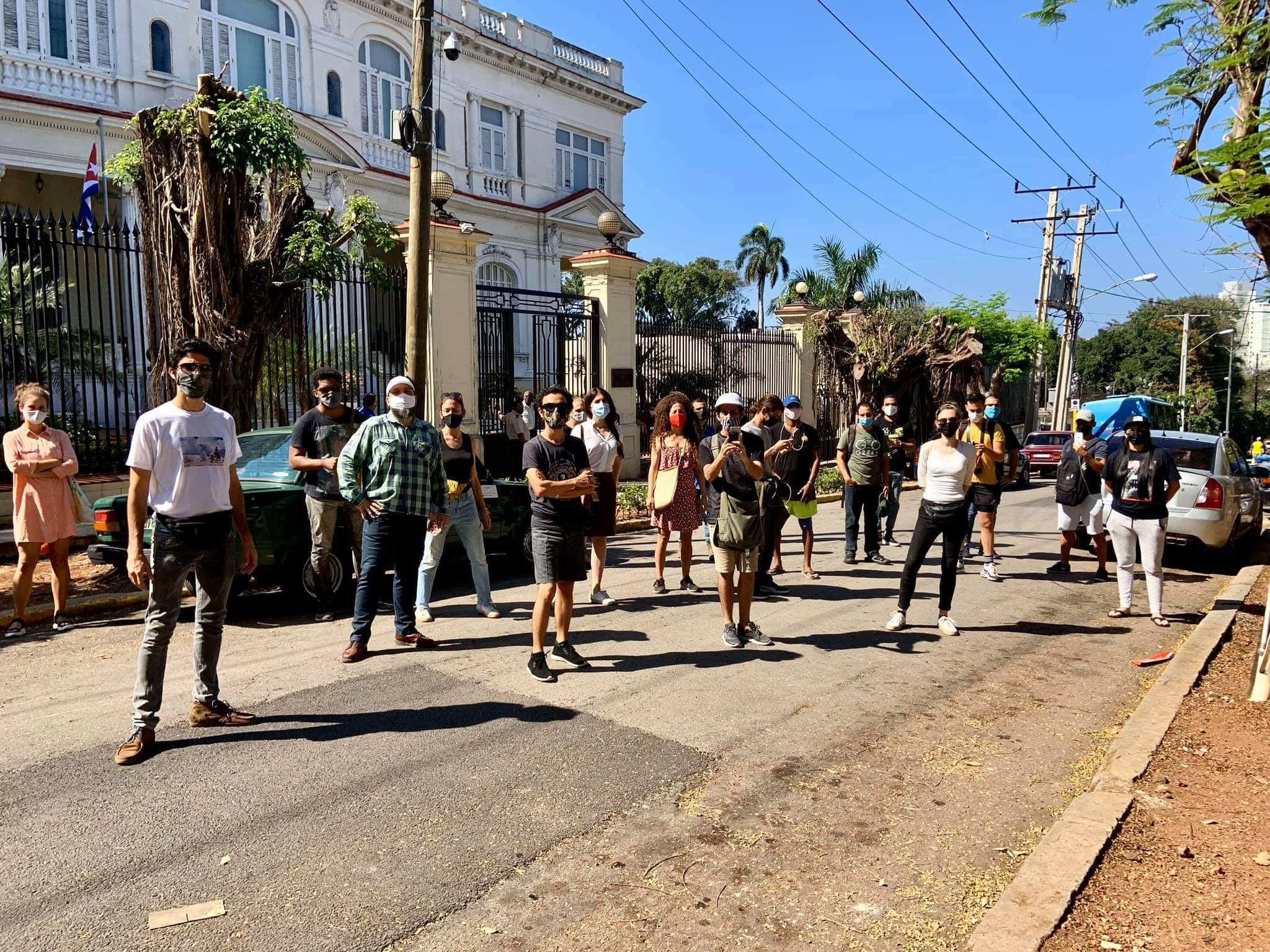 A group of people wearing masks stand spaced apart on a sunny street in front of a large, white building with a gated fence and palm trees.
