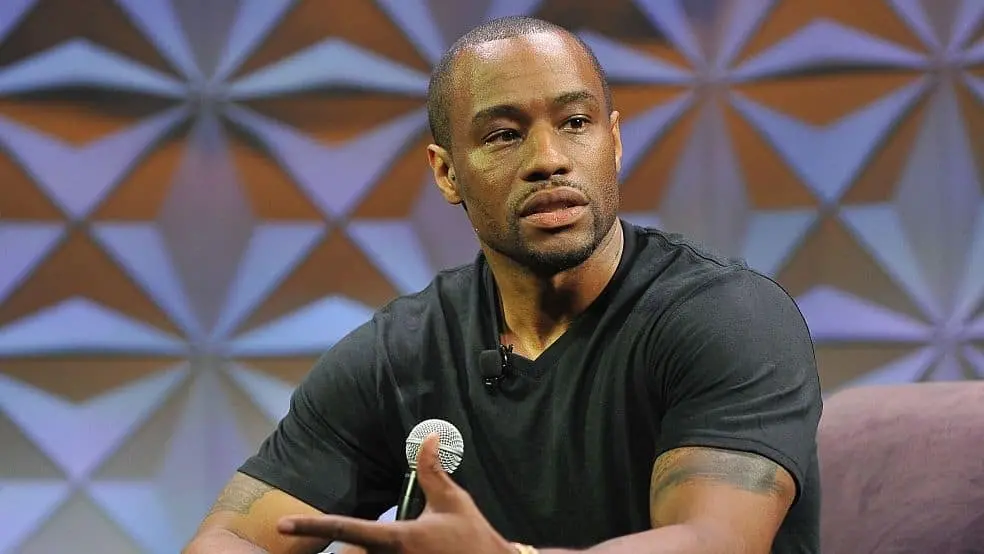A man in a black t-shirt sits on a stage holding a microphone, gesturing with his hand while speaking. The background is a geometric patterned wall in shades of gray and orange.