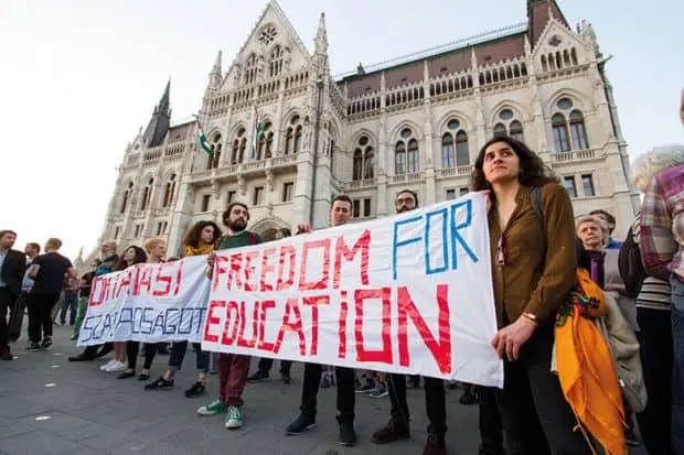 A group of people stand in front of a grand historic building, holding banners that read FREEDOM FOR EDUCATION and OCTA PAST EDUCATION. The mood appears serious and determined.
