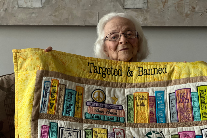 Grace Linn, an elderly woman with white hair and glasses, holds up a colorful quilt with the words Targeted & Banned and fabric books including Harry Potter, Anne Frank, and Fahrenheit 451 sewn onto it.