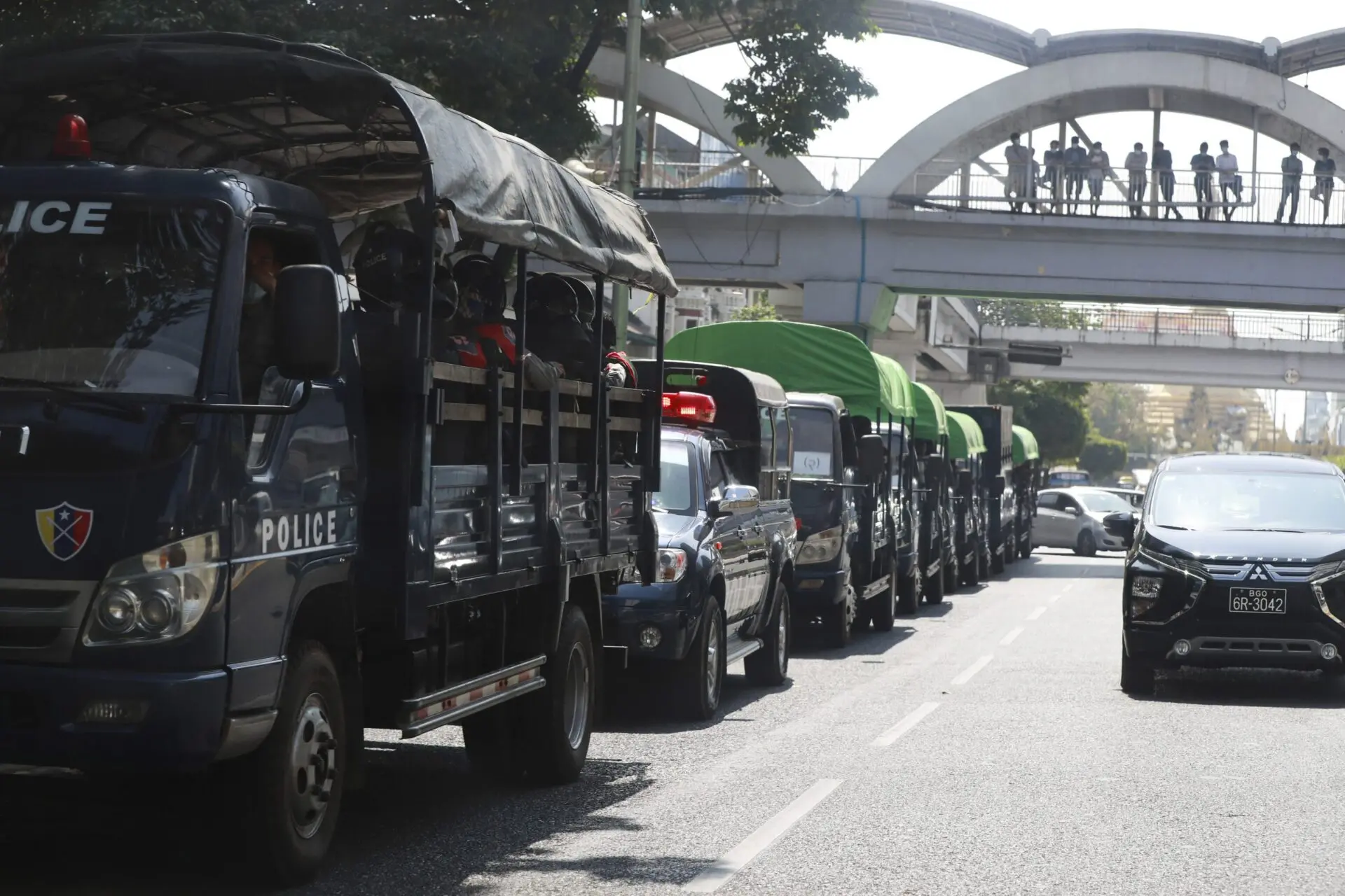A row of police trucks is parked along a city street in daylight, with officers inside. People can be seen standing on a pedestrian bridge above the road.