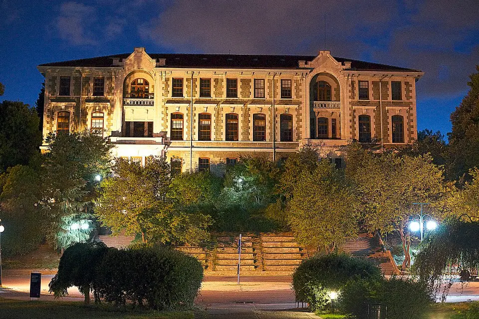 A large, historic building with illuminated windows stands behind trees and stairs at night, under a partly cloudy sky. The scene is softly lit by nearby streetlights.