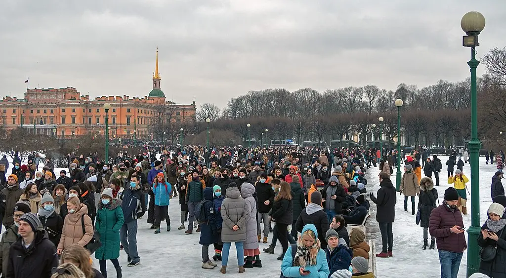 A large crowd of people, many wearing winter coats and some with masks, gathers outdoors in a snowy park with trees and a historic building in the background under a cloudy sky.