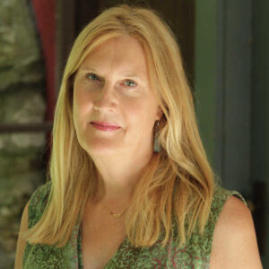 A woman with long blonde hair wearing a green patterned sleeveless top stands indoors, looking at the camera with a gentle expression, as if ready for writer interviews. A blurred background includes a window and greenery.