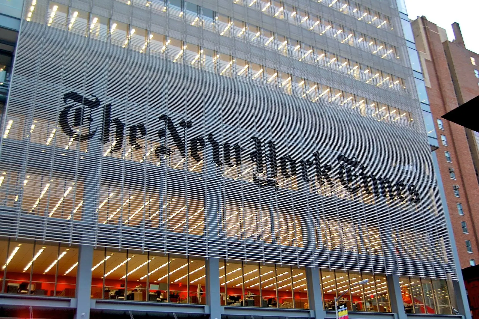 The exterior of The New York Times building features a large sign with the companys name in black lettering, with rows of illuminated office windows visible behind a metal grid façade.