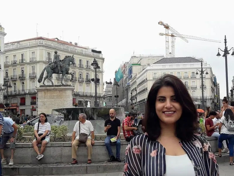 A woman smiles in the foreground of a busy city square, reminiscent of Loujain Al-Hathlouls spirit, with people gathered around a statue of a horse and rider, framed by historic buildings and construction cranes in the background.