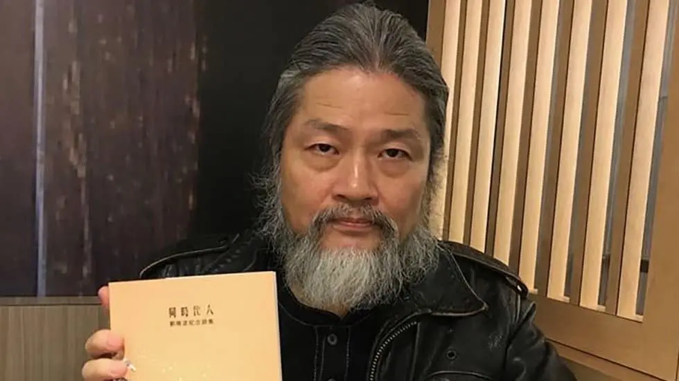 A man with long gray hair and a beard sits indoors, holding up a book with Asian script on the cover. He is wearing a black jacket and looking directly at the camera.