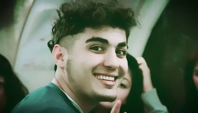 A young man with short, curly dark hair and a trimmed beard smiles at the camera, wearing a green shirt. He is indoors with blurred people and a curved wall in the background.