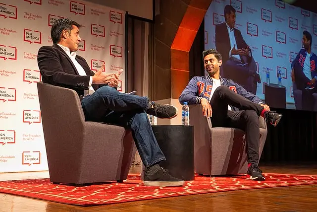 Two men sit and converse on stage in armchairs at a PEN America event, with a screen behind them displaying their image. Both are smiling, engaged in discussion, and the stage has a red patterned carpet.