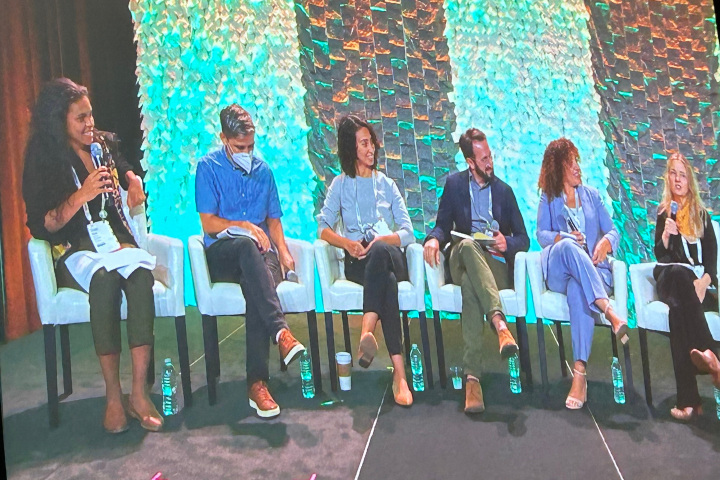 Six people sit on stage in white chairs during a panel discussion. One woman on the far left speaks into a microphone. The background features a textured, illuminated green and orange wall. Bottled water sits at their feet.