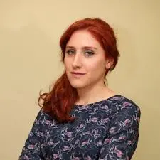 A woman with long red hair and fair skin wears a floral-patterned dark top. She poses against a plain beige background, looking calmly at the camera with her arms crossed.