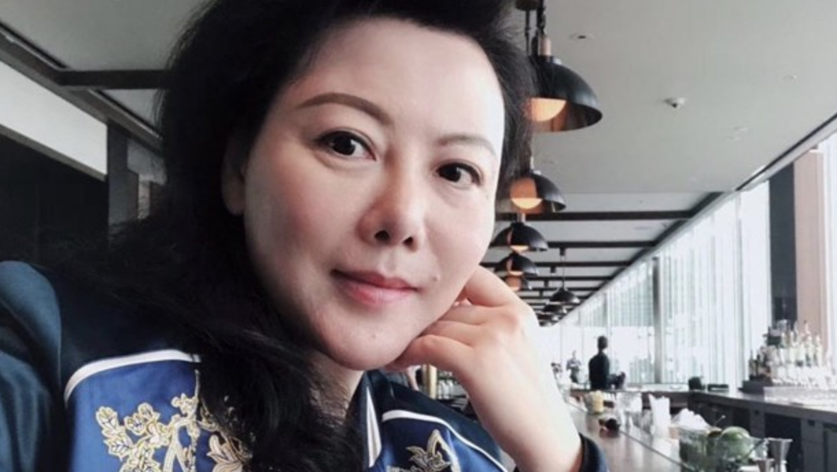 A woman with long dark hair and a blue patterned jacket sits at a bar, resting her head on her hand and smiling. The background shows bar shelves, hanging lights, and large windows.