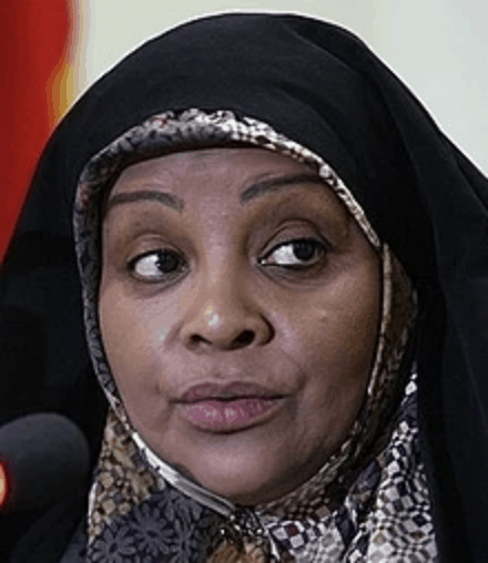 A woman wearing a black hijab and a patterned headscarf speaks into a microphone. The background is plain with a hint of red on the left side.