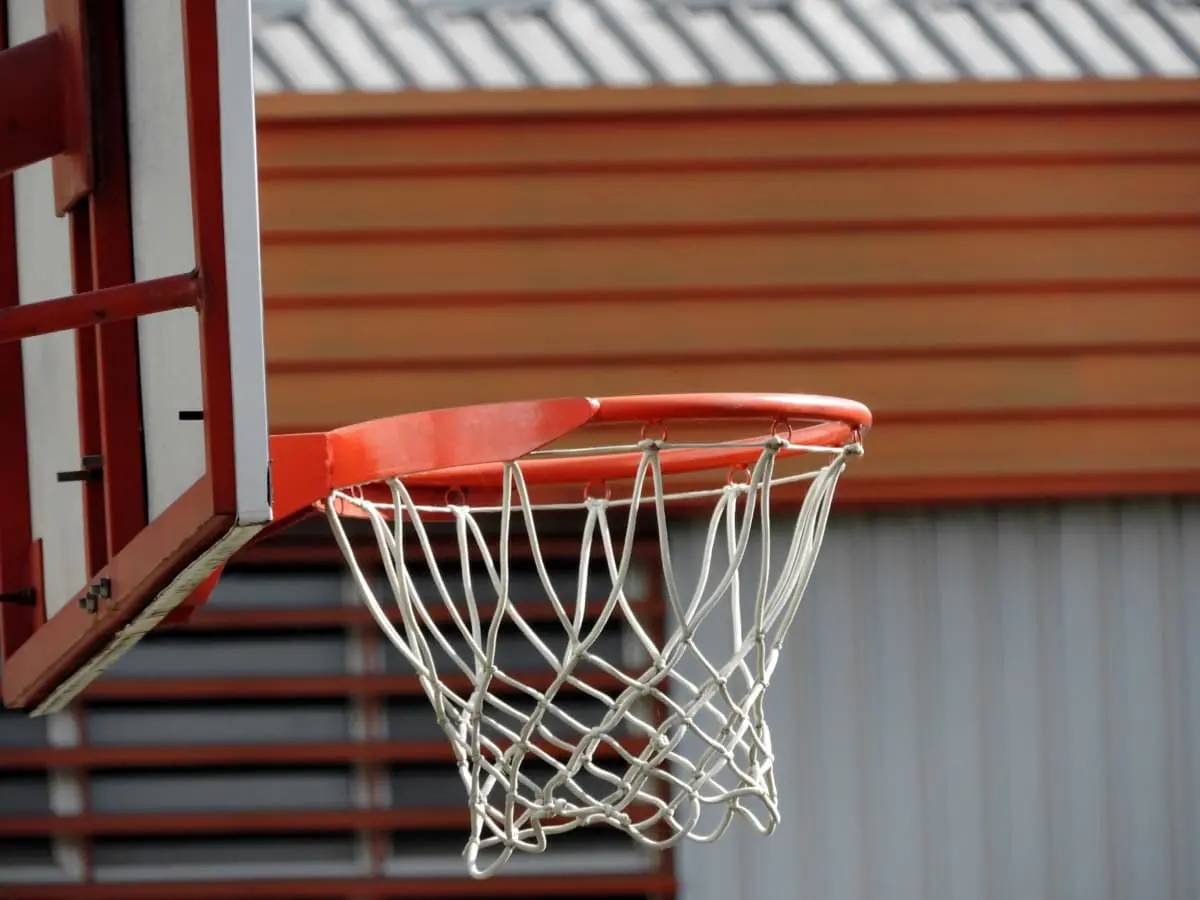 A close-up view of a basketball hoop and net attached to a backboard, with a building featuring orange and gray horizontal siding in the background.