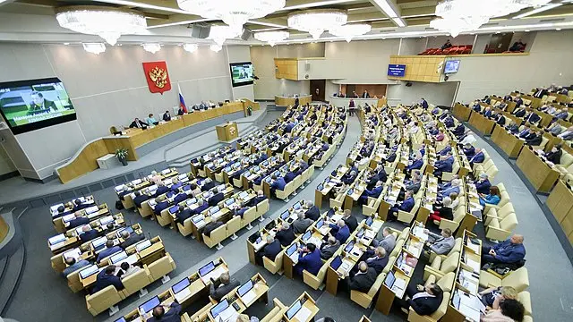 A wide view of a large parliamentary chamber with many seated officials facing a central podium. Multiple rows of desks, large screens, and a Russian coat of arms are visible on the wall.