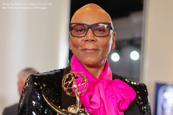 A person wearing a shiny black jacket and bright pink scarf smiles while holding an Emmy award. Channeling RuPaul vibes, they have glasses and are standing indoors.