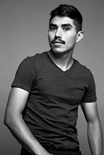 A man with short dark hair and a mustache poses against a plain background, wearing a short-sleeve, dark-colored t-shirt. He looks slightly to the side with a neutral expression.