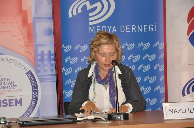 A woman with blonde hair sits at a table, speaking into a microphone at a conference. She is reading from a paper. Behind her are banners with text and logos. A nameplate on the table reads Nazlı Ilıcak.