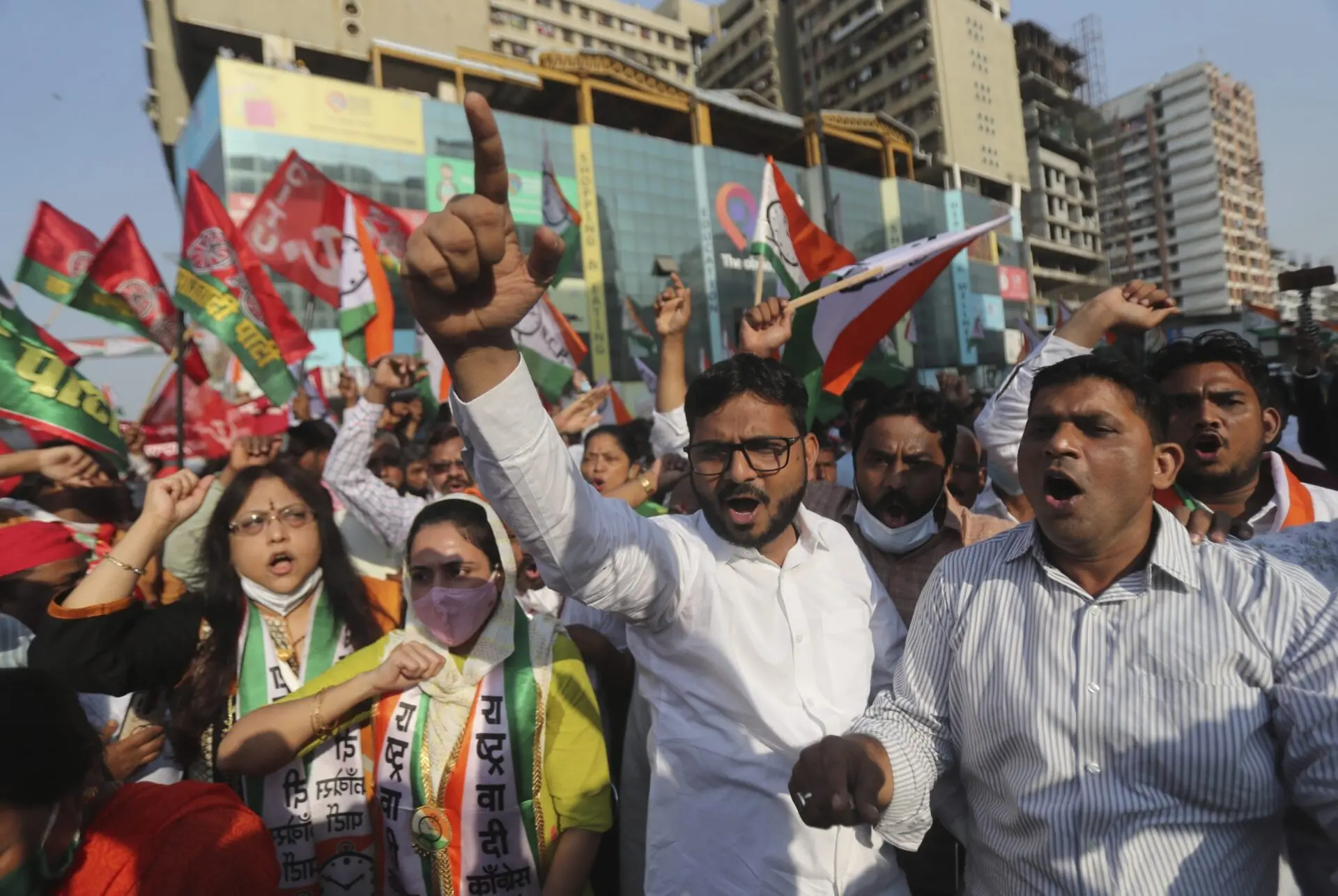 A group of people protest on a city street, raising their fists and chanting. Some hold flags and banners, while others wear masks and protest scarves. Tall buildings and a crowd are visible in the background.