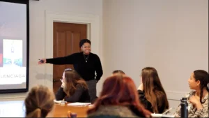 A person standing and smiling while pointing to a projected screen during a presentation on Harvard free speech in a conference room, with several people seated at a table listening attentively.